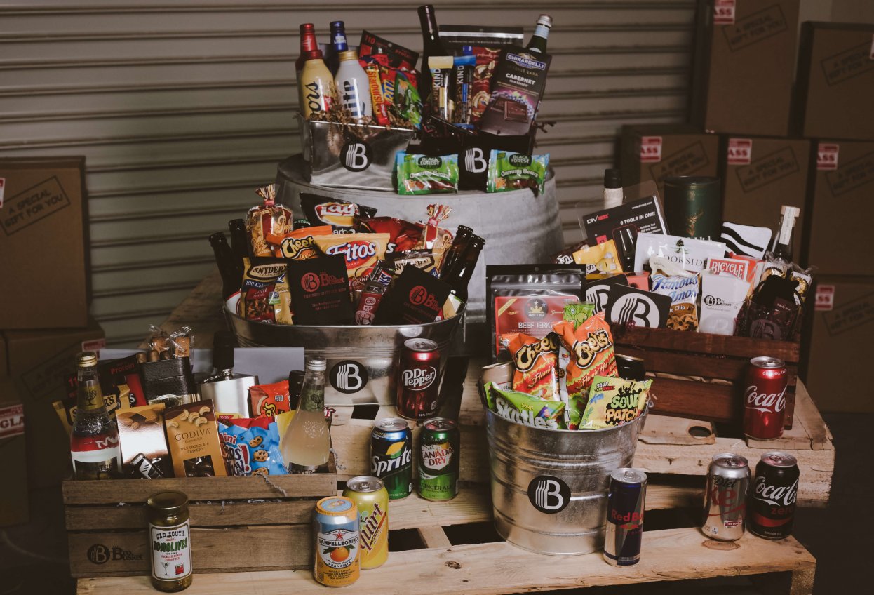 Assorted gift baskets from BroBasket with snacks and beverages, including chips, sodas, and chocolates, displayed on wooden pallets.