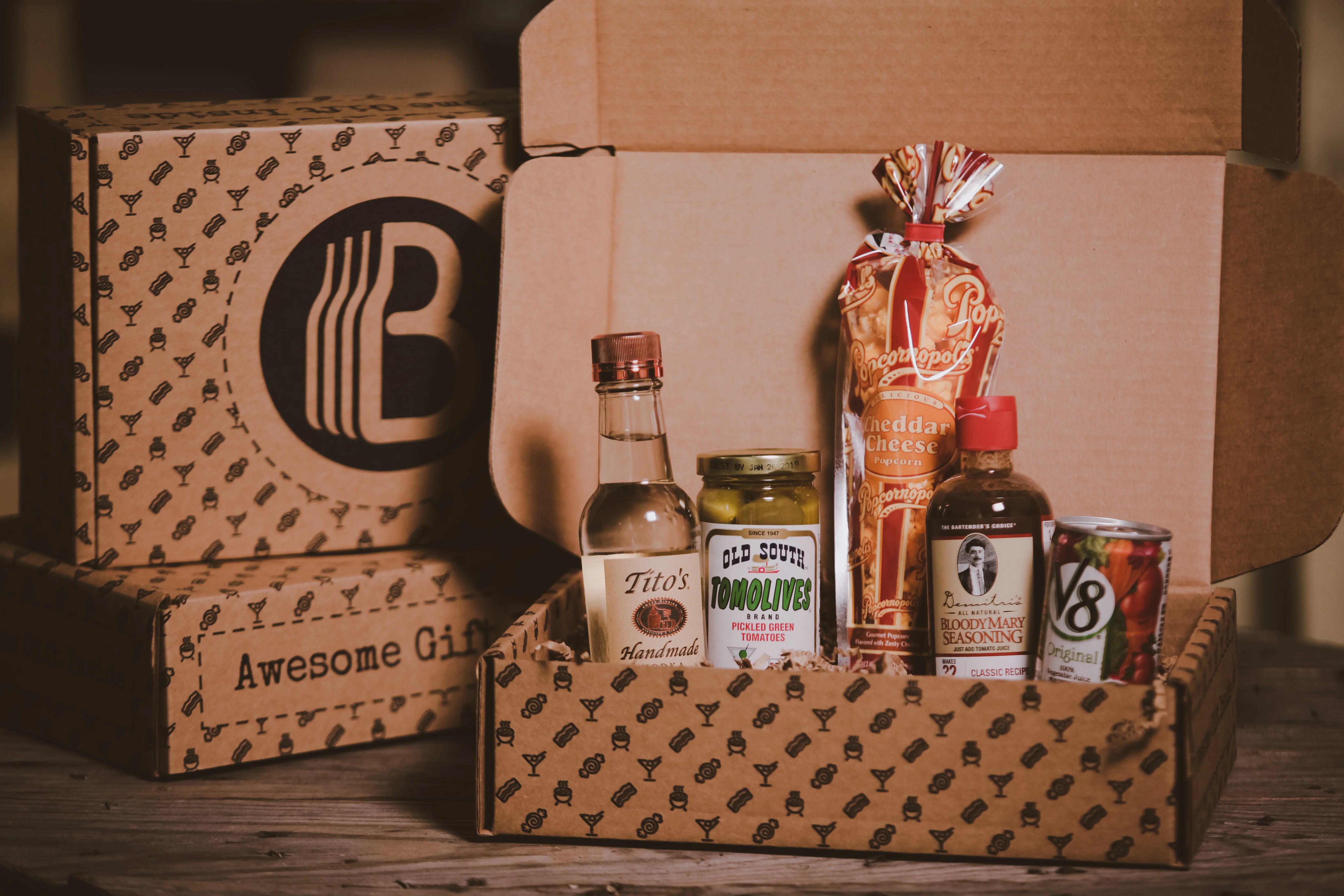 Gift basket from BroBasket containing Tito's vodka, Old South Tomolives, cheddar cheese popcorn, Bloody Mary seasoning, and V8 juice, displayed in branded packaging on a wooden surface.
