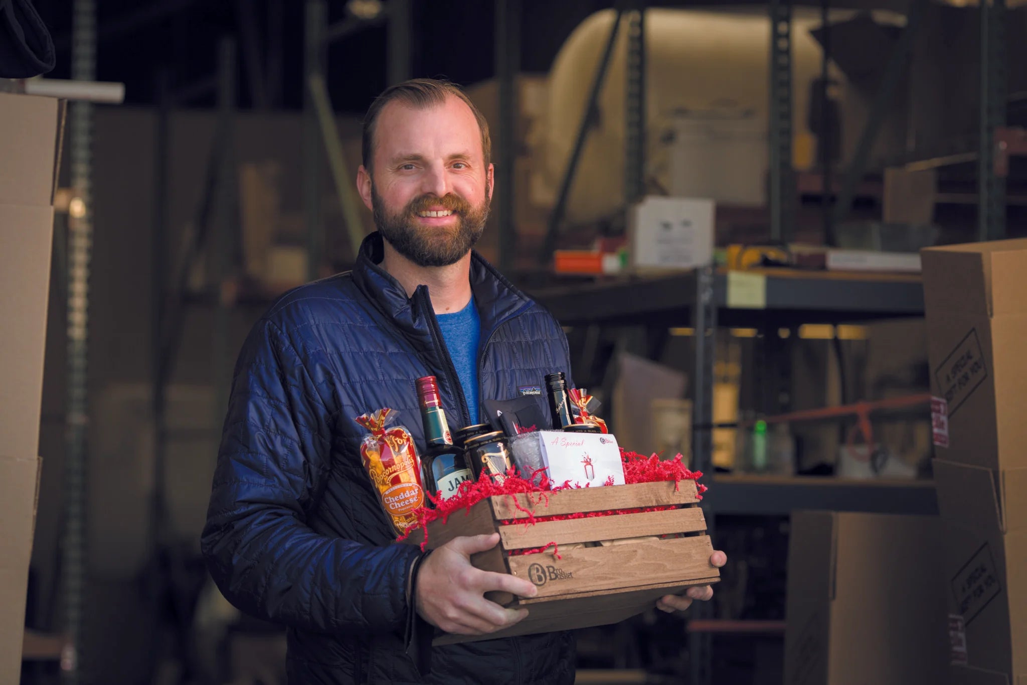 Man in a warehouse holding a wooden BroBasket gift basket filled with bottles and snacks, wearing a blue jacket and smiling.