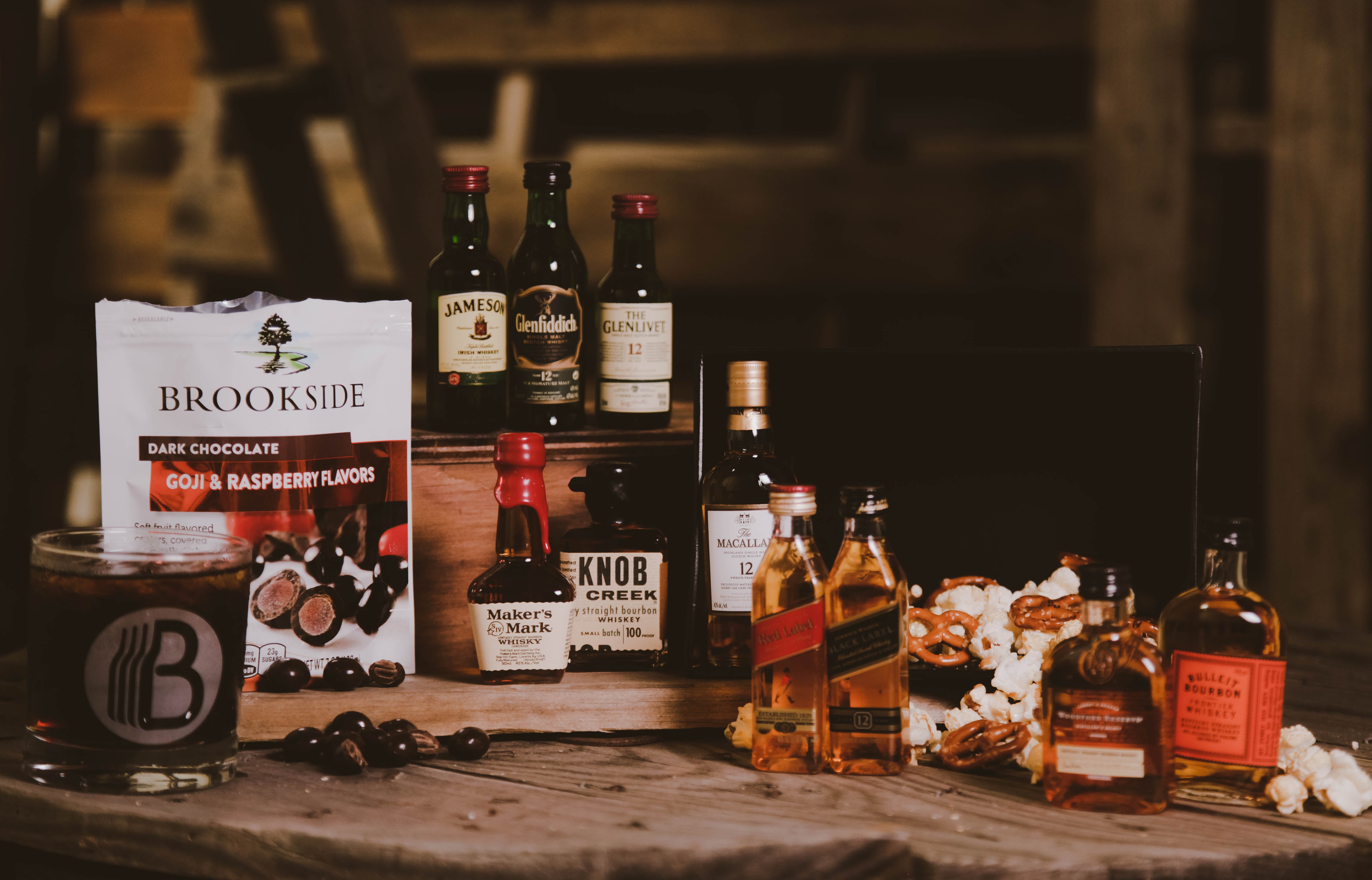 Gift basket from BroBasket including assorted mini bottles of whiskey and bourbon with Brookside dark chocolate, popcorn, and pretzels on a rustic wooden table setting.