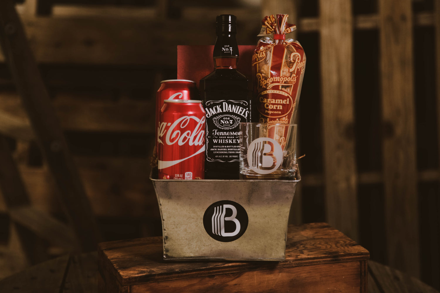 Gift basket from BroBasket containing a bottle of Jack Daniel's whiskey, two Coca-Cola cans, caramel corn popcorn, and a branded glass on a rustic wooden surface.