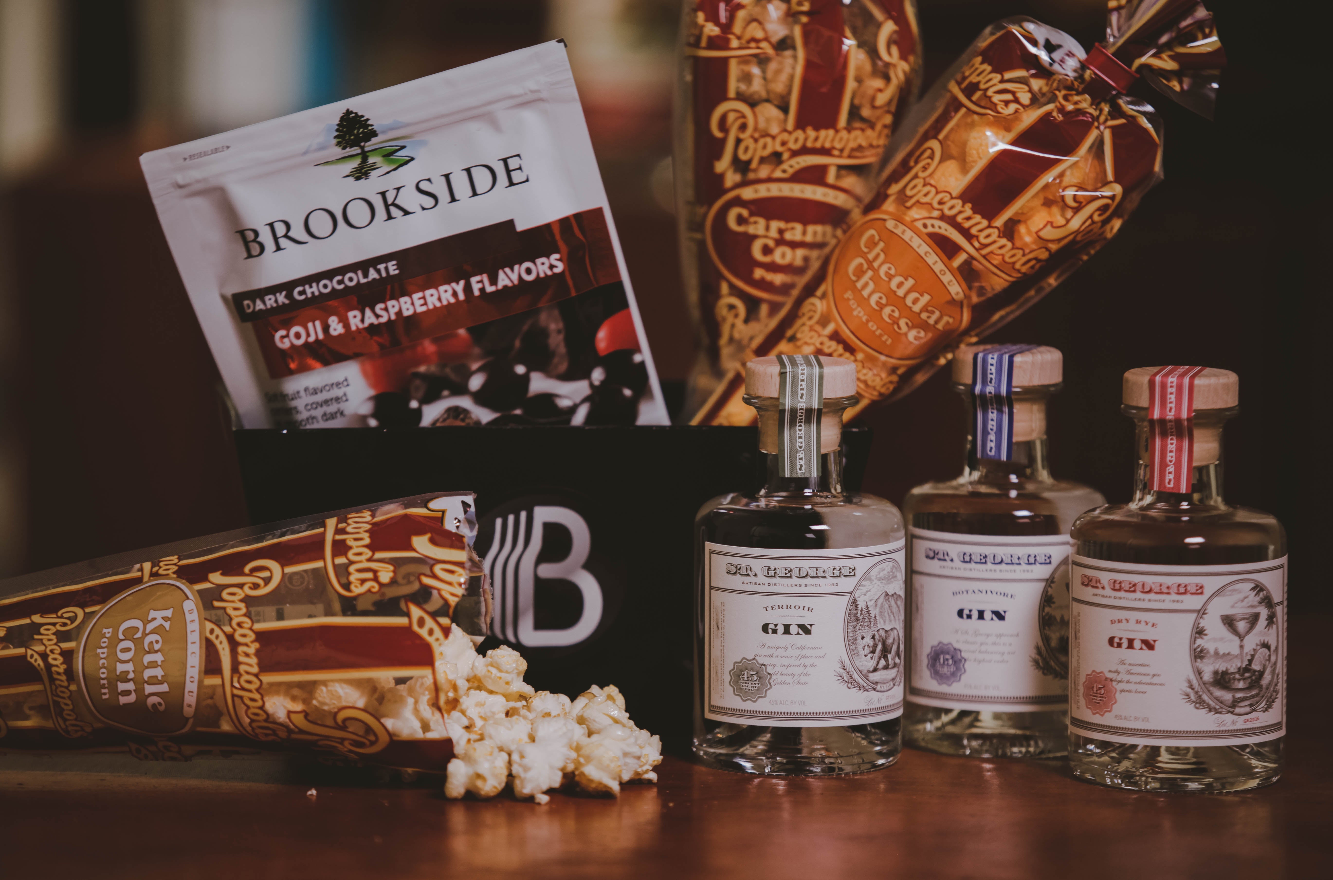 Gift basket from BroBasket including assorted gourmet snacks and gin bottles including Brookside dark chocolate, Popcornopolis popcorn, and St. George gin displayed on a wooden table.