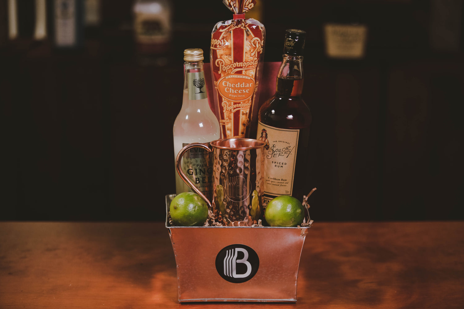 Gift basket from BroBasket with ginger beer, spiced rum, cheddar cheese popcorn, copper mug, and limes on a wooden table.