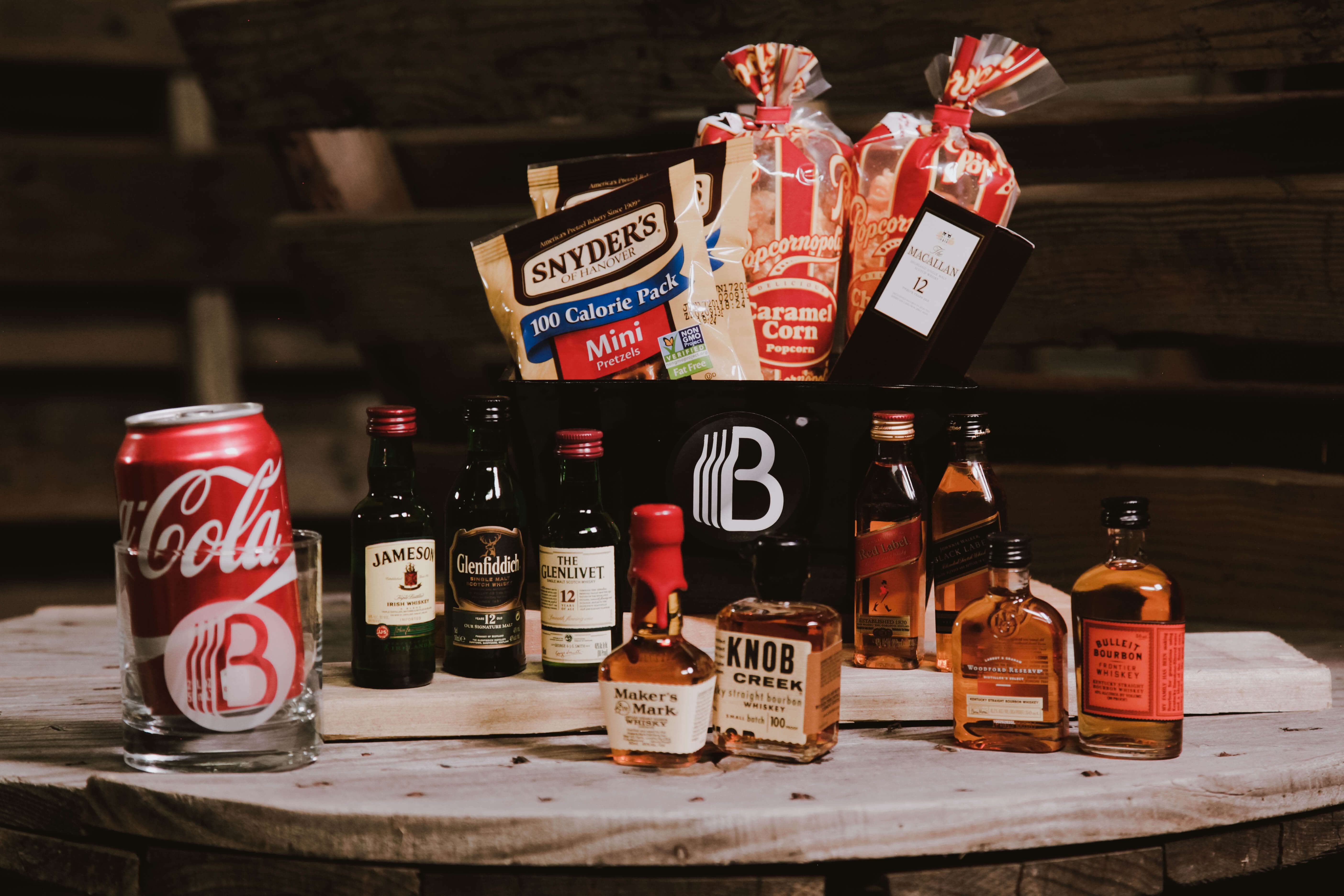 Gift basket from BroBasket including assorted mini liquor bottles, a can of Coca-Cola, pretzels, and caramel corn arranged on a wooden barrel table.